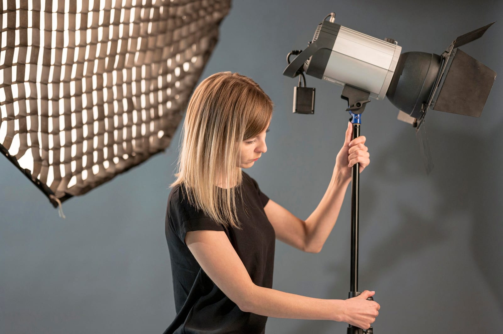 Mulher ajusta a altura de um refletor com bandeiras ao lado de um softbox em estúdio de fundo cinza